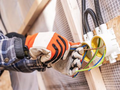 An electrician connects colored wires to a junction box while wearing gloves and working diligently. The setting shows a construction site with wooden frames.